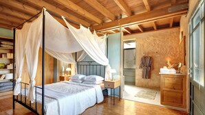 Quarto com cama de dossel, vigas de madeira no teto, mesa de cabeceira com abajur e banheiro com toucador e espelho de madeira.
