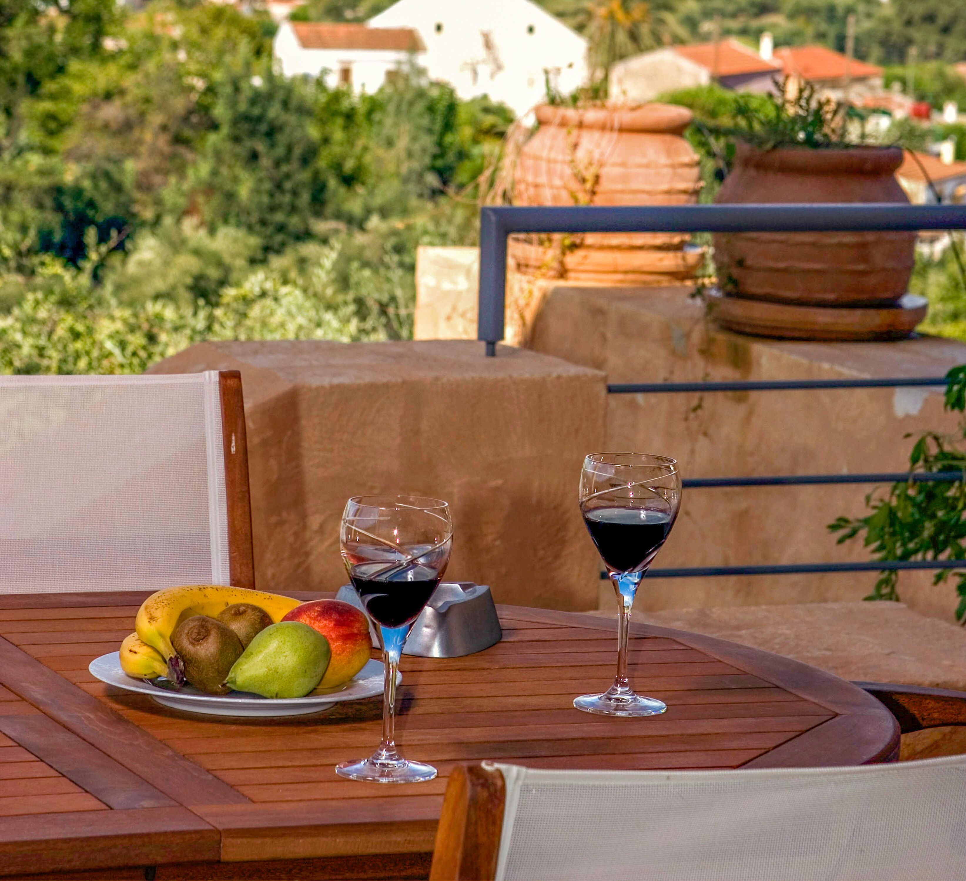 A wooden table with a plate of fruit and two wine glasses on a balcony overlooking a scenic landscape.