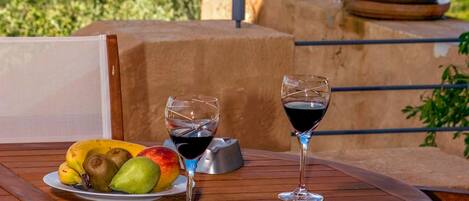 A wooden table with a plate of fruit and two wine glasses on a balcony overlooking a scenic landscape.