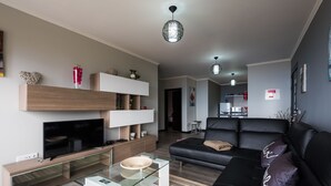 A modern living room with a flat-screen TV, glass coffee table, black leather sofa, and a dining area with a small table and chairs. - Super OCEAN VIEWS - Modern 2 Bed Apartment Stylishly Furnished (Arco da Calheta)