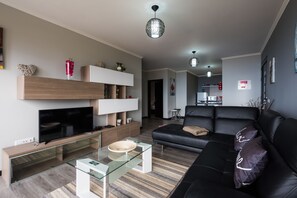A modern living room with a flat-screen TV, glass coffee table, black leather sofa, and a dining area with a small table and chairs.