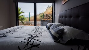 A bedroom with a large bed, a view through a sliding door to an outdoor area with a stone wall and plants. - Super OCEAN VIEWS - Modern 2 Bed Apartment Stylishly Furnished (Arco da Calheta)
