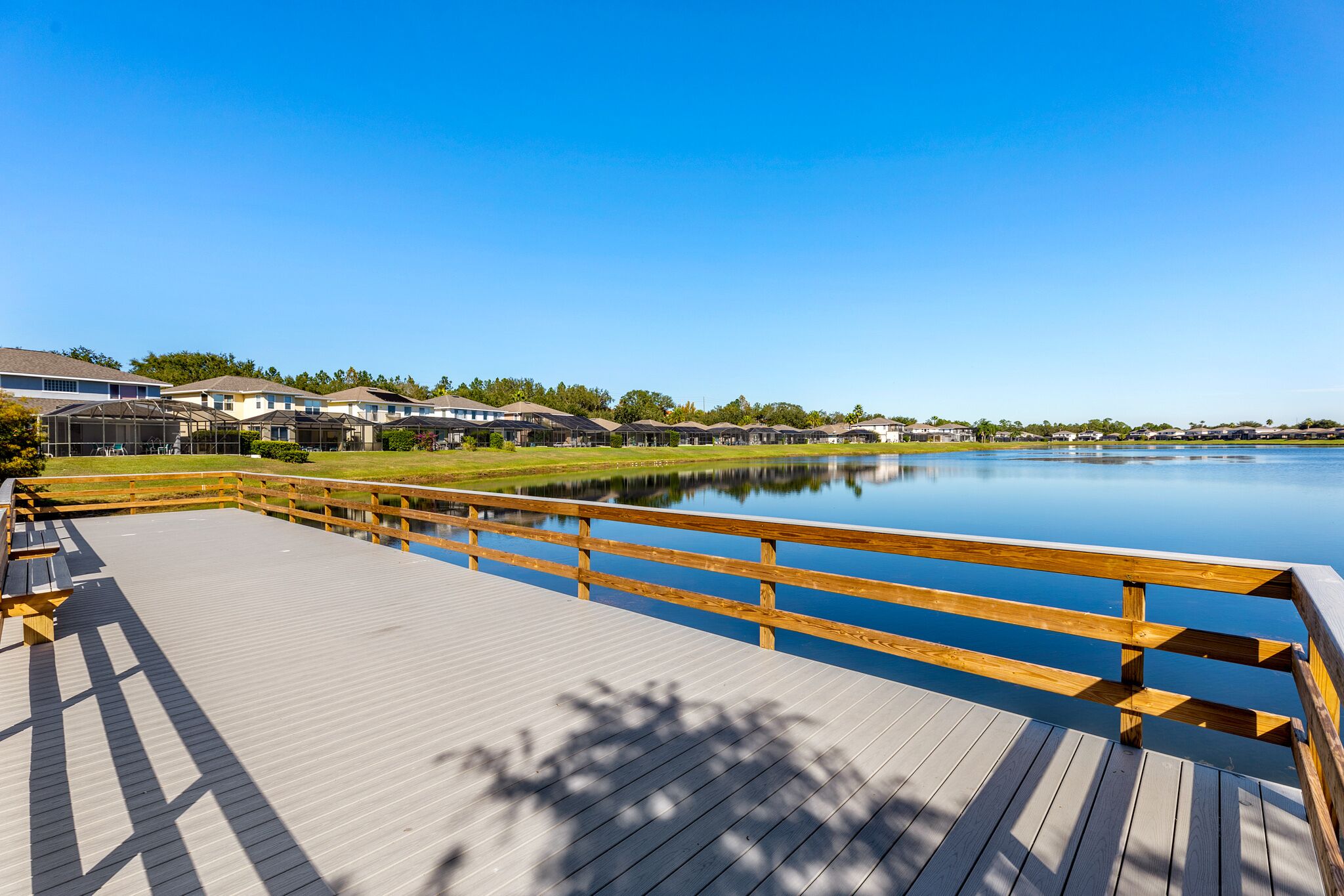 A wooden deck with a wooden railing overlooking a calm lake, surrounded by houses with a clear blue sky above.