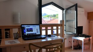 Ein Zimmer mit Schreibtisch aus Holz, einem Laptop, einem Notizbuch und einem Stuhl. Ein Fenster mit Jalousien, ein kleiner Tisch mit Fernseher und eine Wand-Klimaanlage sind ebenfalls vorhanden.