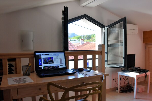 Una habitación con un escritorio de madera, una computadora portátil, un cuaderno y una silla. Hay una ventana con persianas, una mesita con televisor y un aire acondicionado de pared.