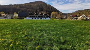 Ein weißes Gebäude mit schwarzem Dach, umgeben von einem grünen Feld mit gelben Blumen, vor der Kulisse eines bewaldeten Hügels unter einem teils bewölkten Himmel.