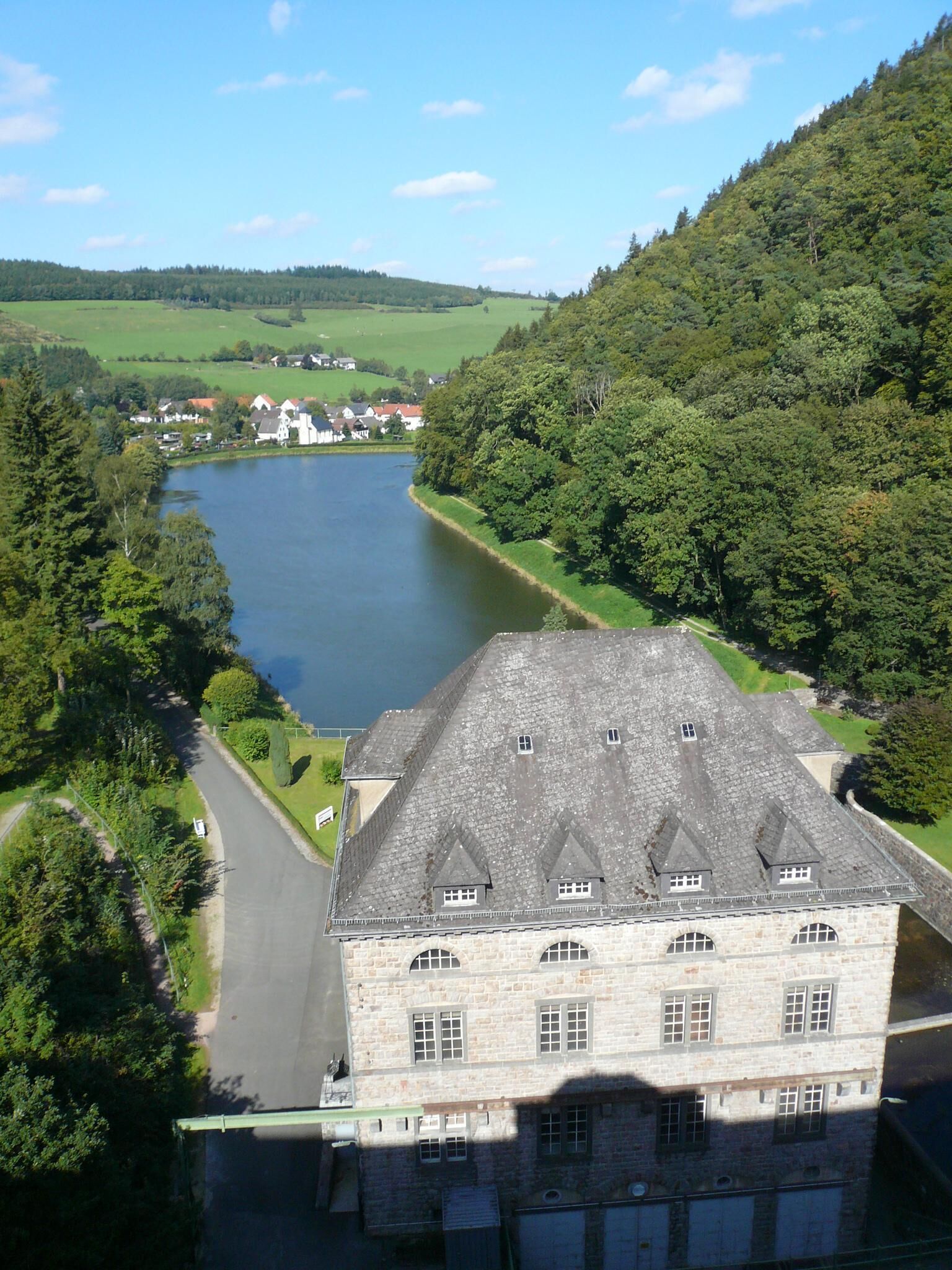 Ein großes Steinhaus mit einem steilen Dach, an einem Fluss gelegen, mit einer kleinen Stadt im Hintergrund.