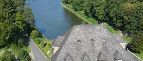 A large stone building with a steep roof, situated beside a river with a small town in the background.