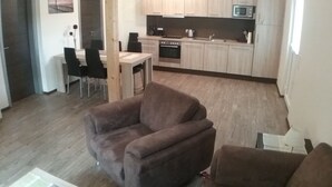 A modern living room with a brown sofa, a dining area with a table and chairs, and a kitchen with light wooden cabinets and a microwave. - Welcome to our apartments of Villa Wald-Eck (Apartment 2)  (Ückeritz)
