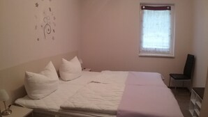 A bedroom with a bed, pillows, a bedside table, a chair, a window with curtains, and a wall decoration. - Welcome to our apartments of Villa Wald-Eck (Apartment 2)  (Ückeritz)