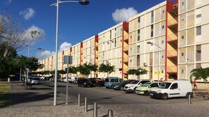 Un immeuble résidentiel à plusieurs étages, doté d’une façade uniforme, de balcons et d’un espace de stationnement avec des voitures.