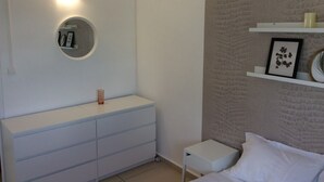 Une chambre avec une commode blanche, un miroir rond, un lit avec une literie blanche et une étagère avec des photos encadrées.