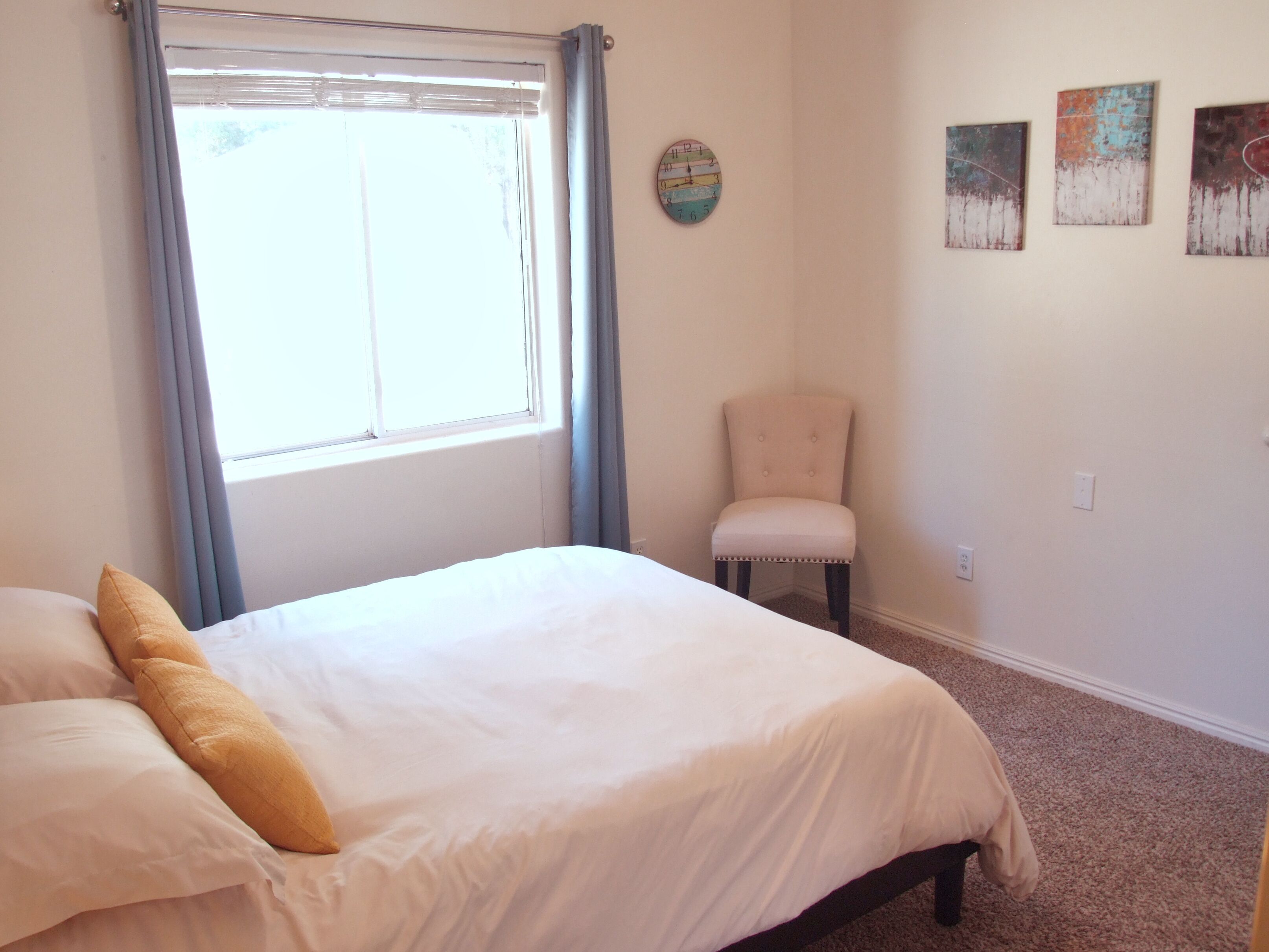 A bedroom with a bed, a chair, a window with curtains, and three wall art pieces.