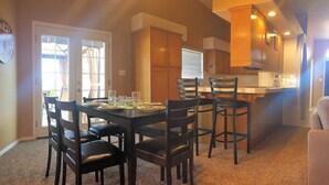 Un coin repas avec une table dressée pour six personnes, entourée de chaises, dans une pièce dotée de meubles en bois et où l’on aperçoit une cuisine au fond.