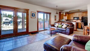 A spacious living room with a leather sofa and a coffee table, a kitchen with wooden cabinets, and large sliding glass doors leading to a deck. - Super Ski Pad - True Ski-In/Ski-Out Retreat. Private Hot Tub. Walk to Adventure (Breckenridge)