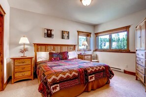 A bedroom with a wooden headboard, a large bed with a colorful quilt, two bedside tables with lamps, a dresser, and a window with a view of trees. - Super Ski Pad - True Ski-In/Ski-Out Retreat. Private Hot Tub. Walk to Adventure (Breckenridge)