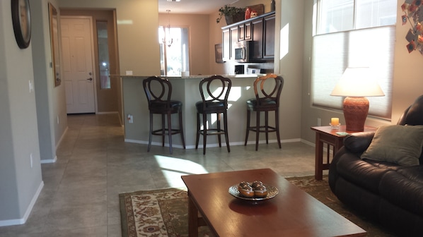 A living room with a brown leather sofa, a wooden coffee table with a decorative plate, and a patterned rug. There is a kitchen area with a bar and three bar stools.
