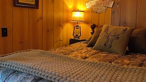 A cozy room with wooden walls, a framed picture, a lamp, a sign reading "Bunkhouse Retreats," and a bed with a camouflage-patterned blanket. - Adorable Mountain Getaway (Shaver Lake)