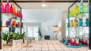 A modern bathroom with glass shelves displaying colorful bottles and plants, a tiled sink area, and a visible window. - Best Views! Golden Girls themed House. Pool and Spa! (Palm Springs)