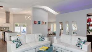A modern living room with white sofas, a glass coffee table, and a kitchen in the background. - Best Views! Golden Girls themed House. Pool and Spa! (Palm Springs)