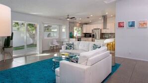 A modern living room with a white sofa, a glass coffee table, and a blue rug. There is a kitchen area with white cabinets and a stainless steel refrigerator. - Best Views! Golden Girls themed House. Pool and Spa! (Palm Springs)