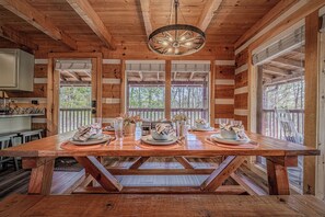 Une table de salle à manger en bois, dressée avec des assiettes, des bols et des couverts, entourée de fenêtres donnant sur un paysage arboré.