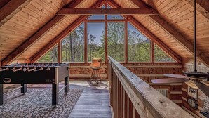 An attic room with a wooden ceiling, a foosball table, a chair, and a large window offering a view of trees. - Hot tub. Views. Multicade & Foosball! (Sevierville)