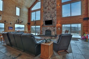 A spacious living area with a stone fireplace, leather sofas, and large windows offering views of the outdoors.