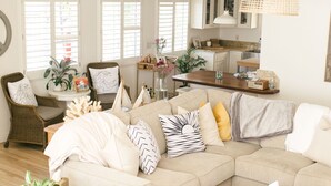 A living room with a sectional sofa, a round ottoman, a wicker chair, and a small side table with a plant. - The Family Beach House  (San Diego)