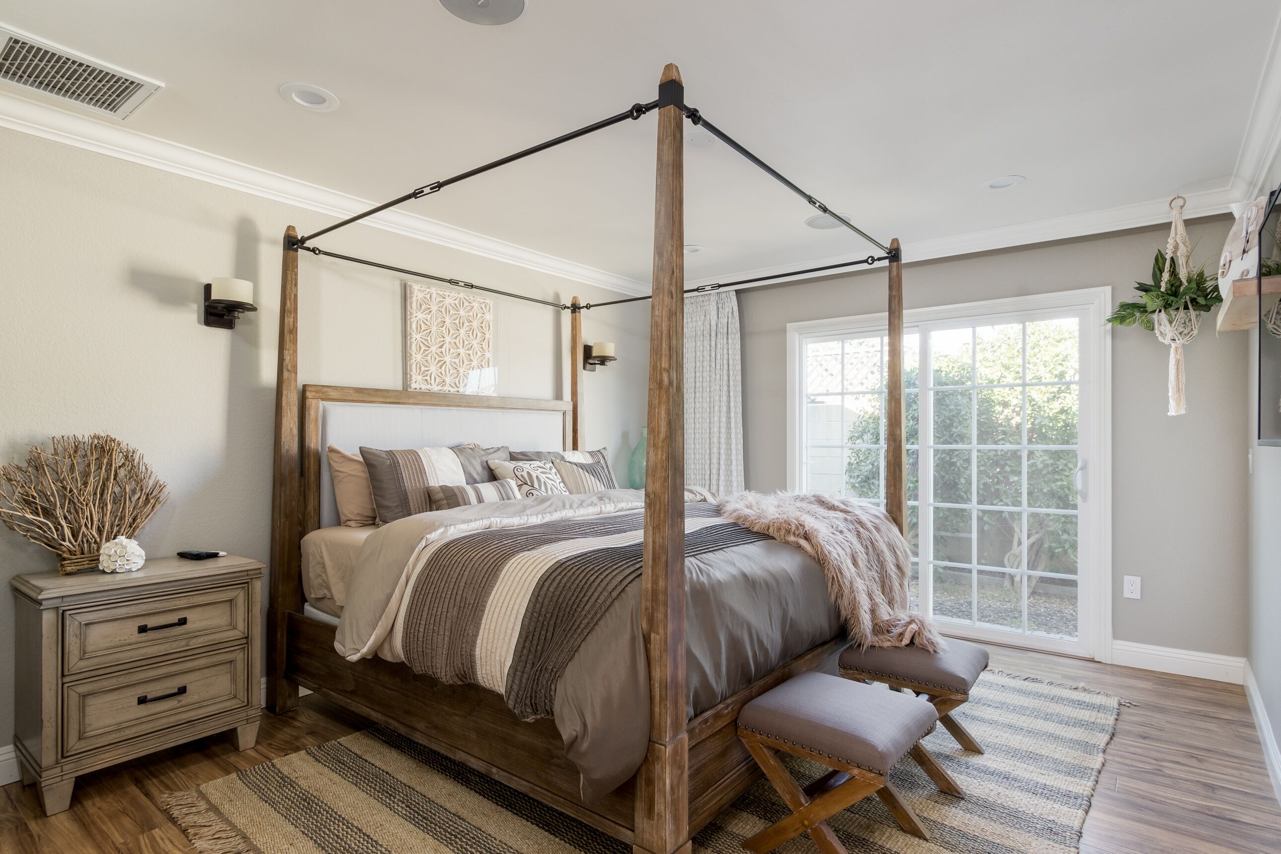 A four-poster canopy bed with a wooden frame, a bedside table with drawers, a bench at the foot of the bed, and a large window with a view of greenery outside.