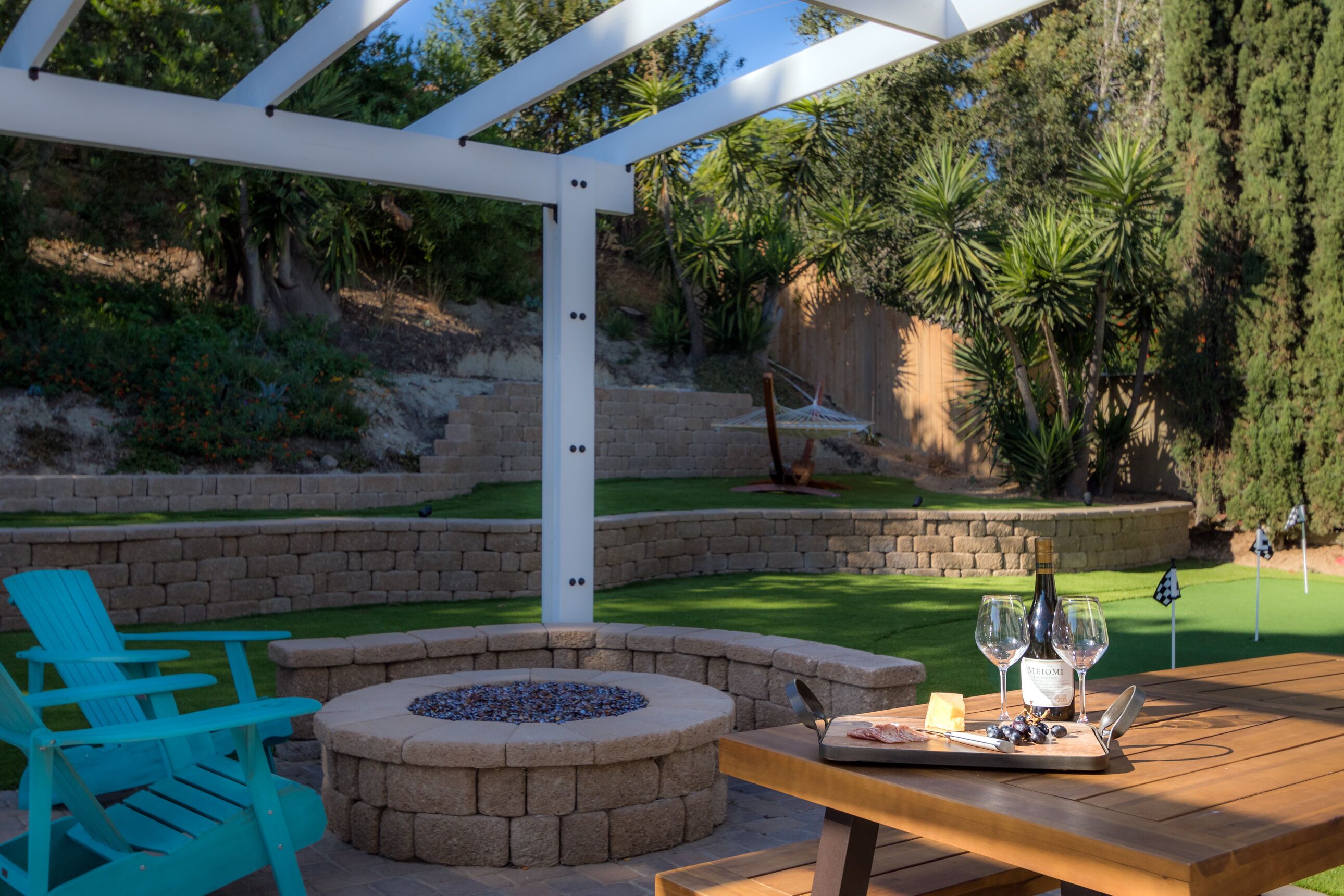 A patio area with a wooden table, a stone fire pit, and a white pergola structure.