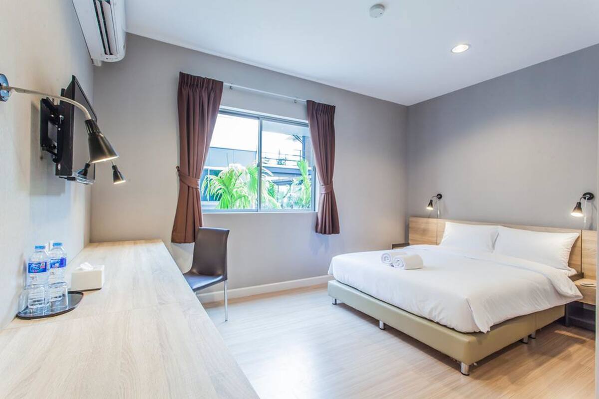 A hotel room with a bed, bedside tables, a desk with water bottles, a chair, a window with curtains, and an air conditioning unit.