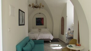 A narrow hallway leading to a bedroom with a bed, a teal sofa, a small table with fruit, and a painting on the wall. - Legatia (Jerusalem)