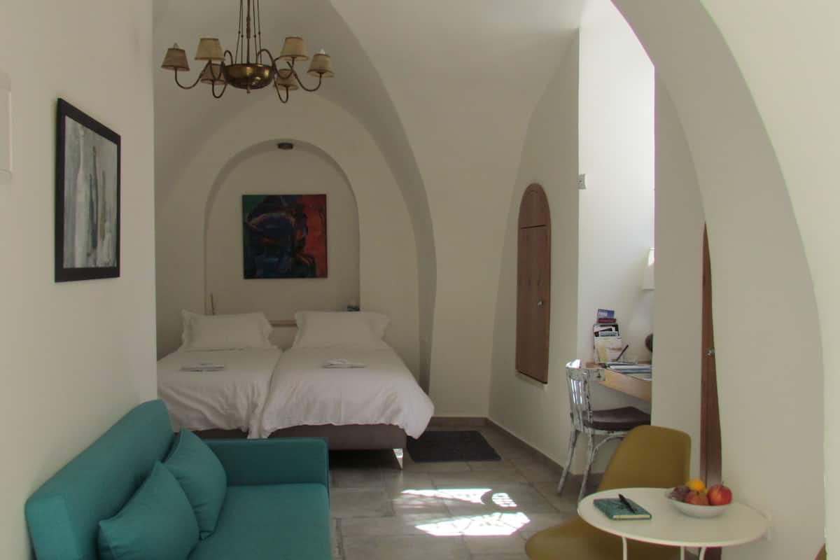 A narrow hallway leading to a bedroom with a bed, a teal sofa, a small table with fruit, and a painting on the wall.