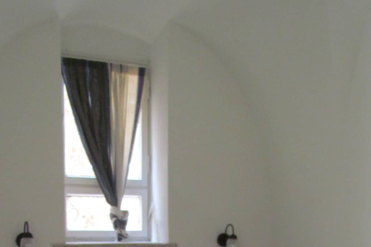 A neatly made bed with white linens, a window with curtains, and two wall-mounted nightstands.