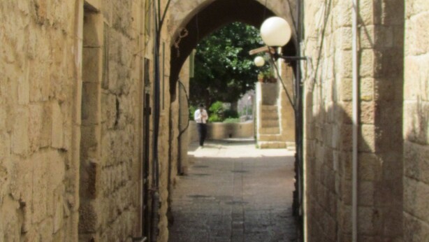 A narrow stone pathway leading to an arched doorway, flanked by buildings with visible windows and satellite dishes. - Legatia (Jerusalem)