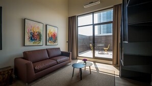 A modern living room with a brown leather sofa, two abstract paintings, a small round table, and a large sliding glass door leading to an outdoor area with yellow chairs. - La Cappella Hotel (Haifa)