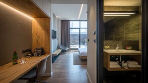 A modern hotel room with a wooden desk, a bed, a bathroom with a large mirror, and a view of the outside. - La Cappella Hotel (Haifa)