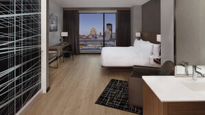 A hotel room with a large bed, a desk, a chair, a desk lamp, a bathroom with a sink and mirror, and a view of the city skyline through large windows.