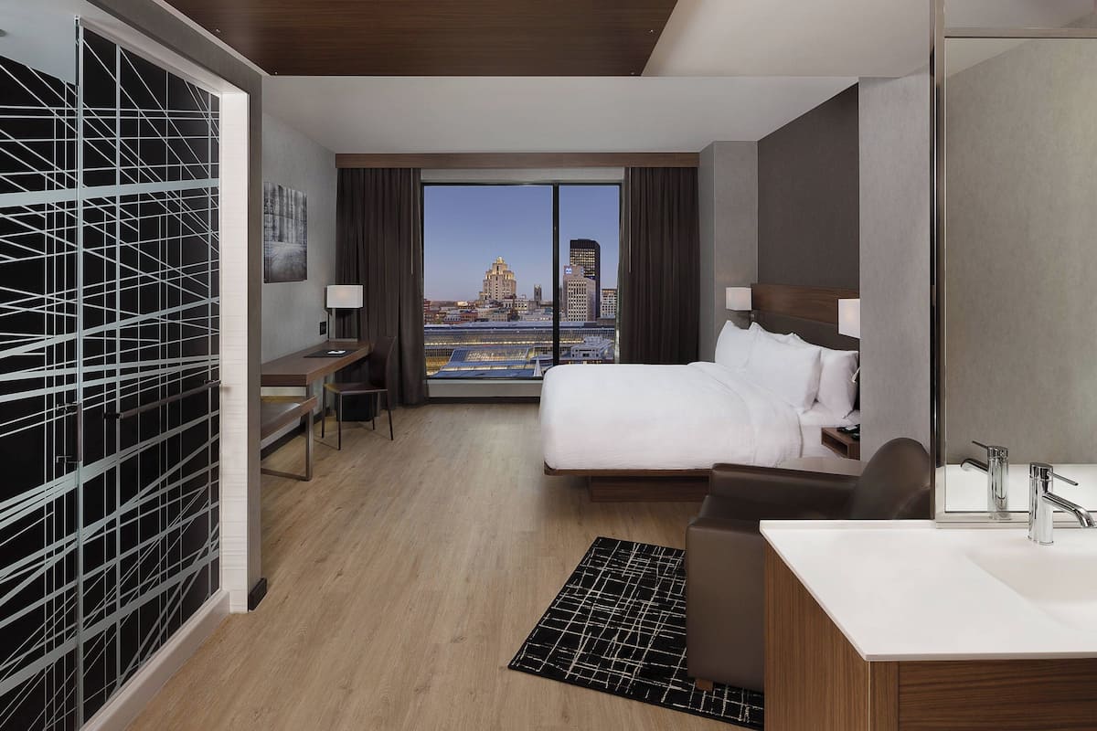 A hotel room with a large bed, a desk, a chair, a desk lamp, a bathroom with a sink and mirror, and a view of the city skyline through large windows.