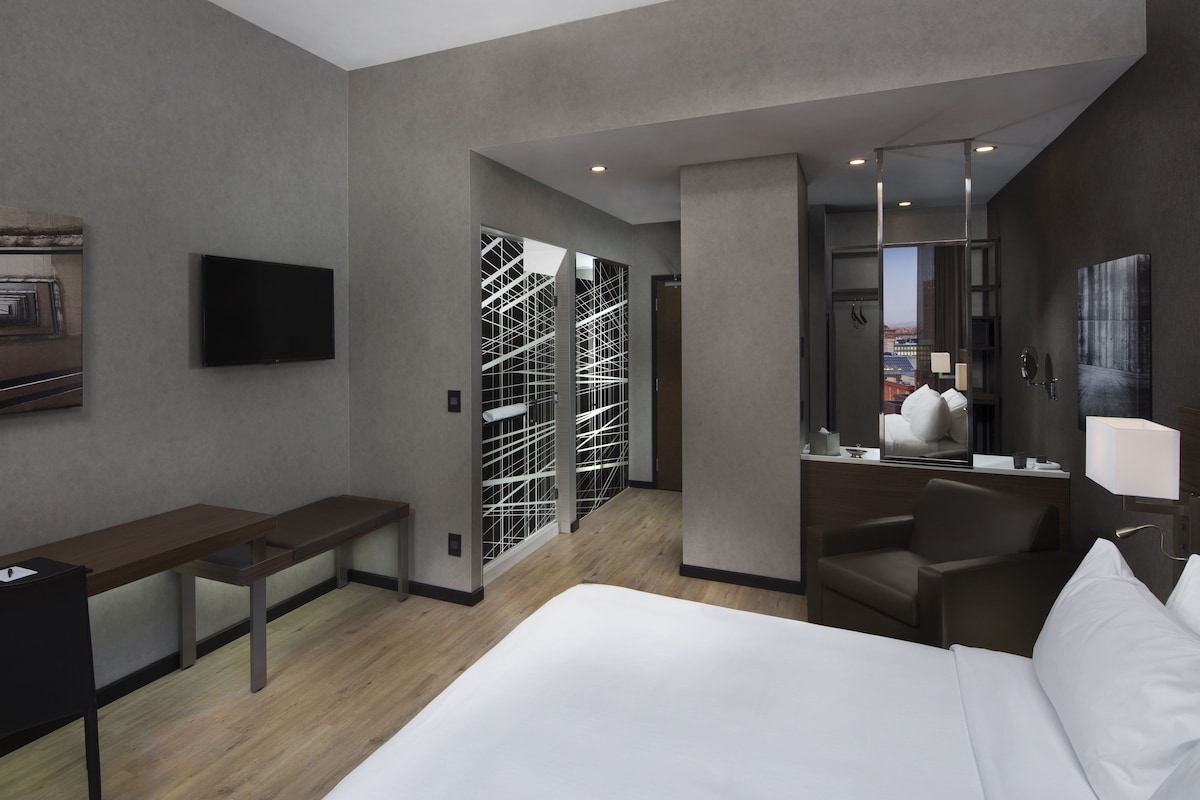 Une chambre d’hôtel moderne, dotée d’un grand lit, d’un bureau, d’une chaise, d’une télévision, d’un miroir et offrant une vue sur la ville par la fenêtre.