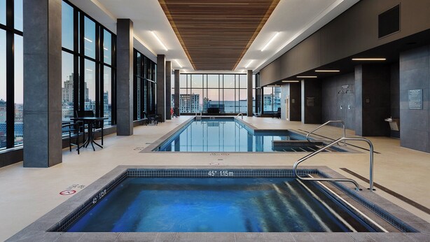 A modern indoor swimming pool with clear glass walls, a wooden ceiling, and a city view.