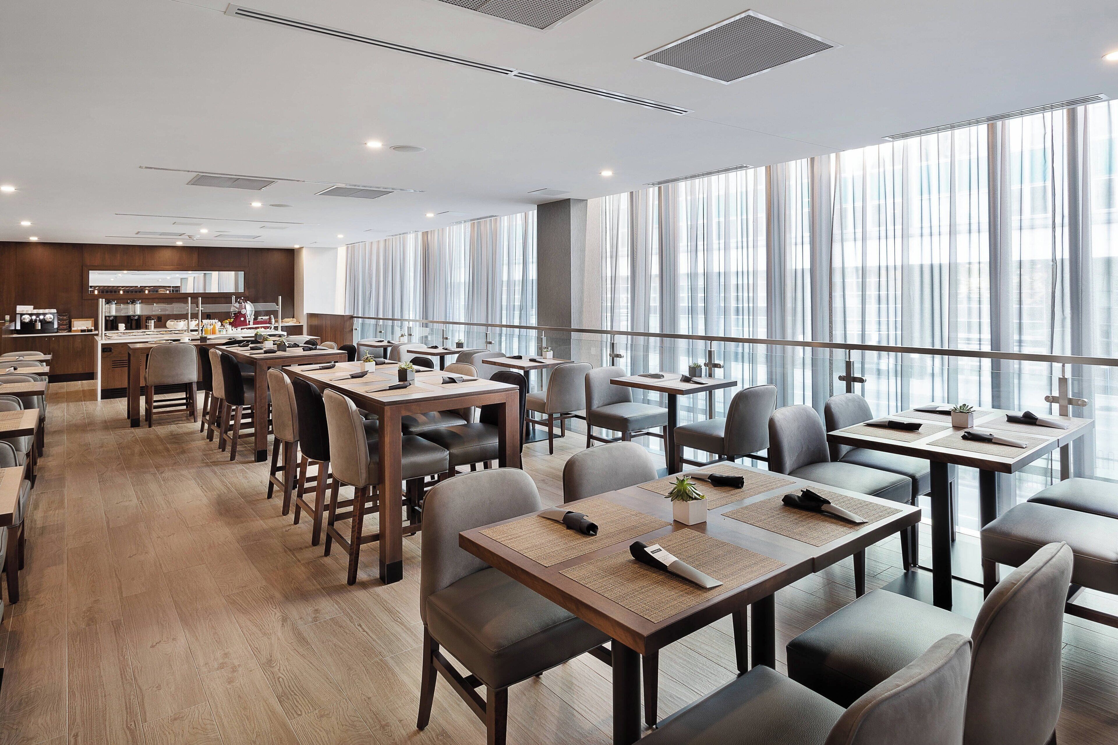 Un espace repas moderne avec des tables et des chaises en bois, de grandes fenêtres et une cuisine en arrière-plan.