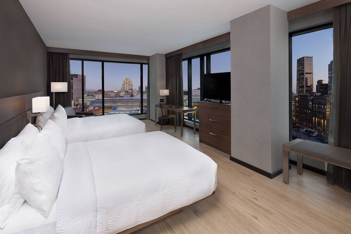 A hotel room with a large bed, a desk, a TV, and a view of the cityscape.