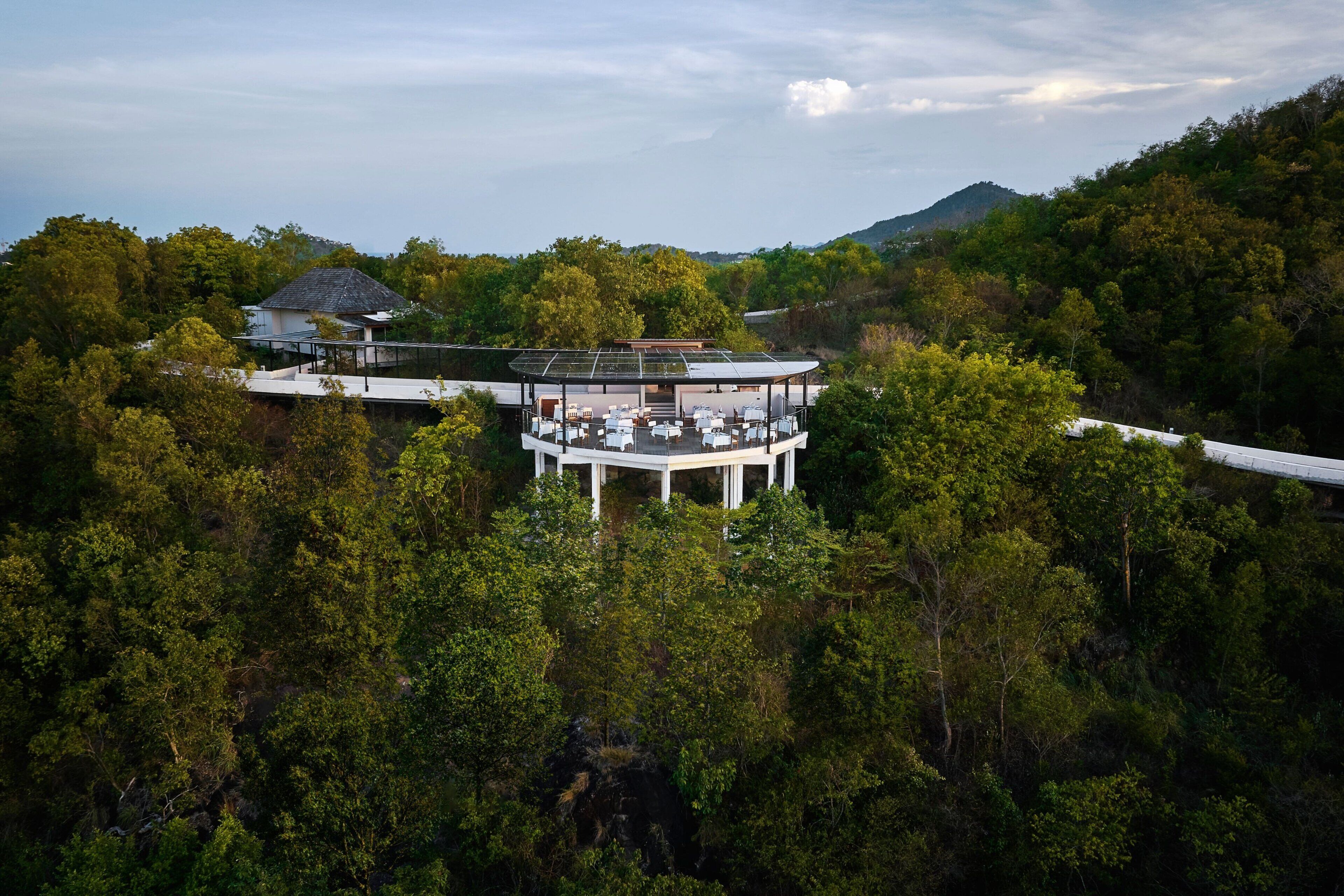 A modern, multi-level building with a glass roof, surrounded by dense forest and hills.