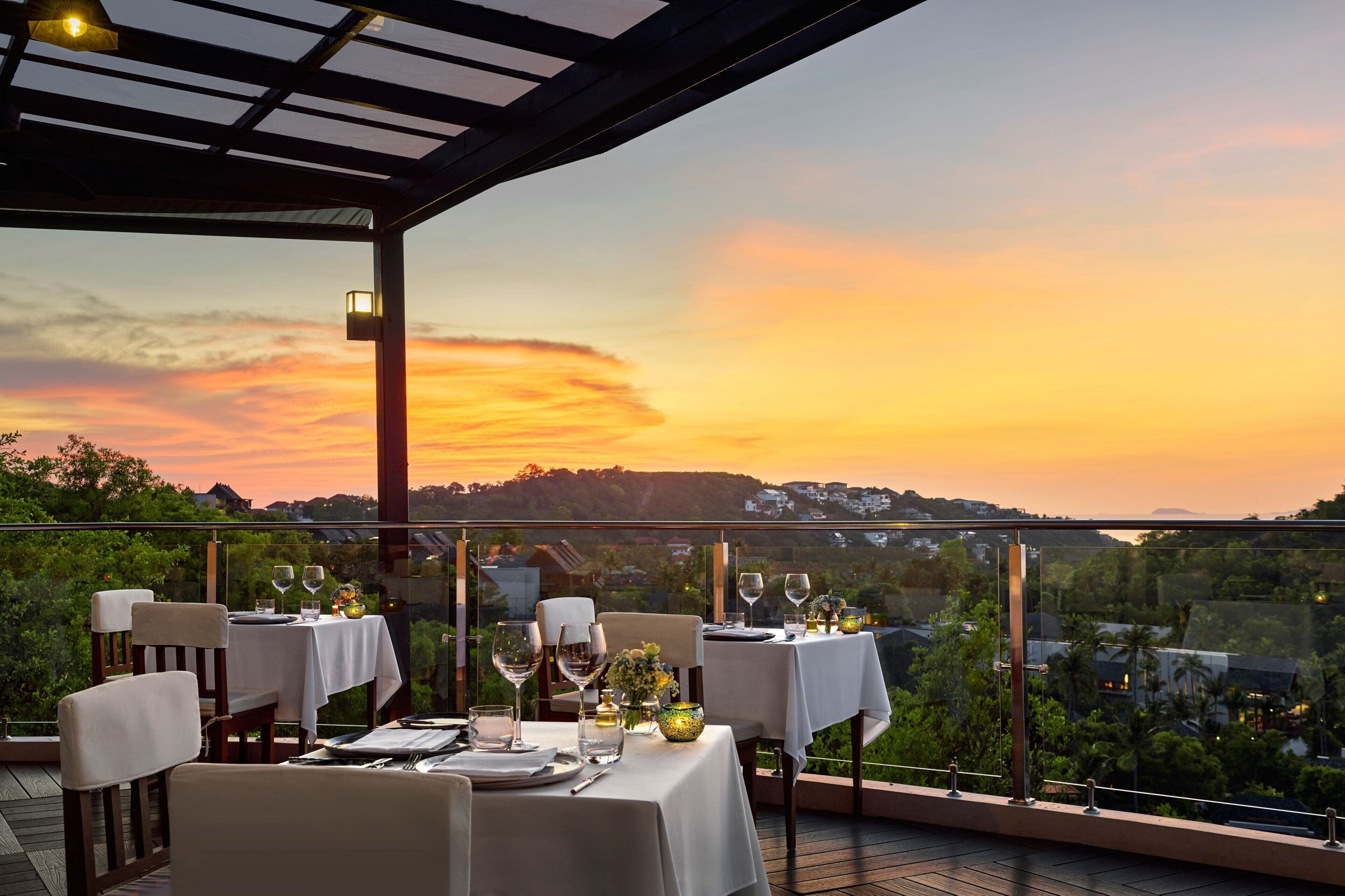 Un espacio de comedor al aire libre con mesas preparadas para dos, con vista a un paisaje escénico de atardecer.