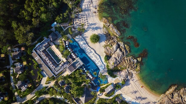 Ett flygbildsbild av ett semesterområde med en stor swimmingpool, strand och en klippig kustlinje.
