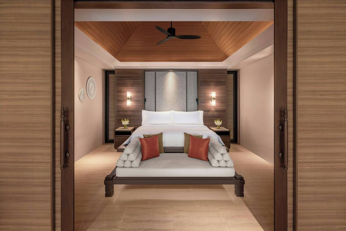 Un dormitorio moderno con techo de madera, una cama con ropa de cama blanca y almohadas rojas, dos mesitas de noche con lámparas y un amplio ventanal con persianas.