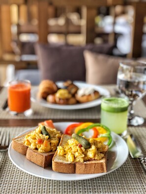 A breakfast setting with toast topped with scrambled eggs and vegetables, a plate of croissants, a glass of orange juice, and a green smoothie on a woven table mat.