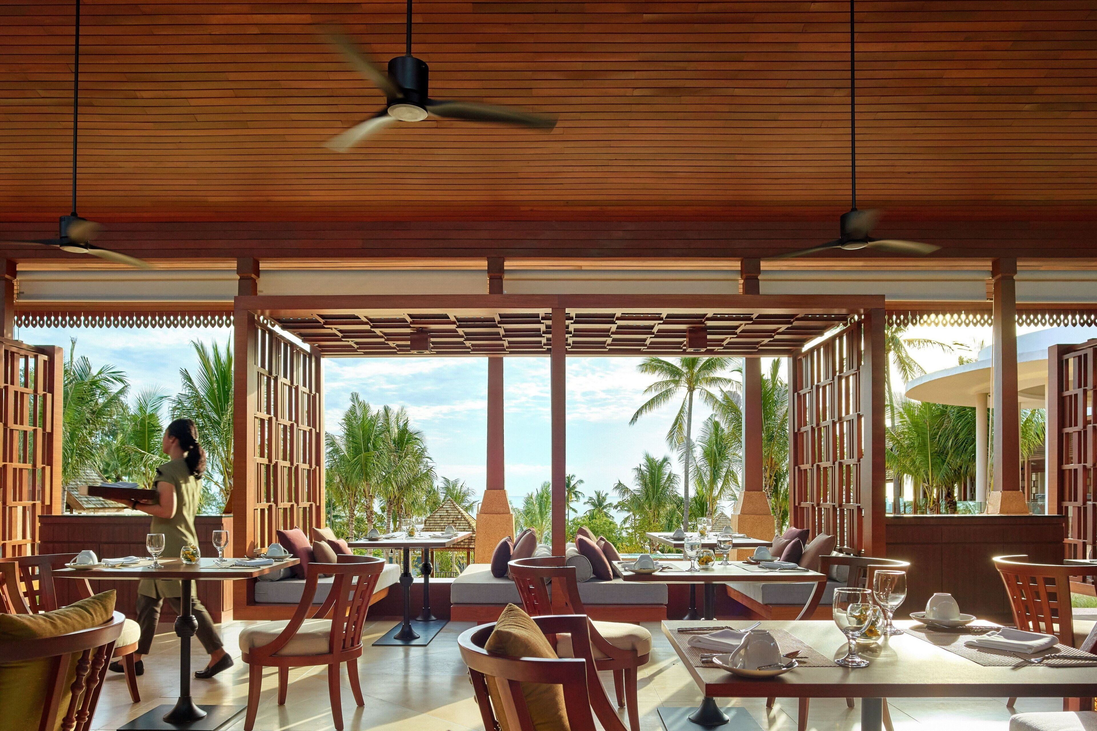 An outdoor dining area with wooden furniture and tables, a waiter carrying a tray, and a view of palm trees and the sea.